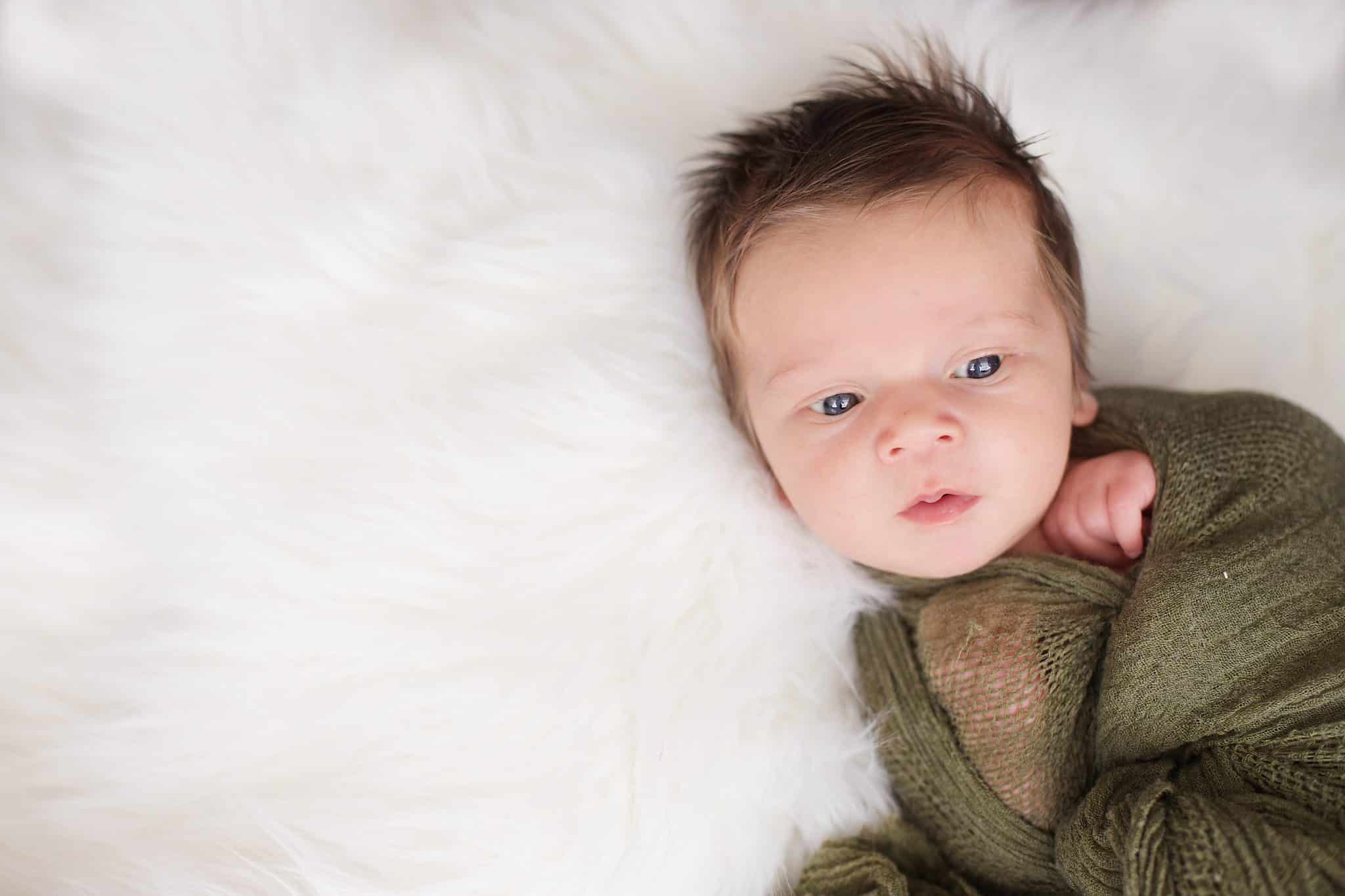 baby boy wide awake wrapped in an olive green wrap on white fur, eyes open