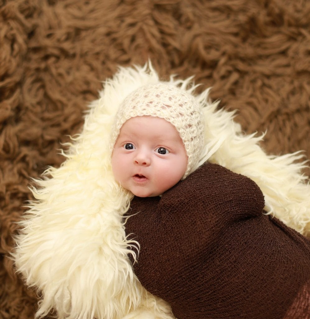 Newborn wrapped in brown, white bonnet, on brown fur 