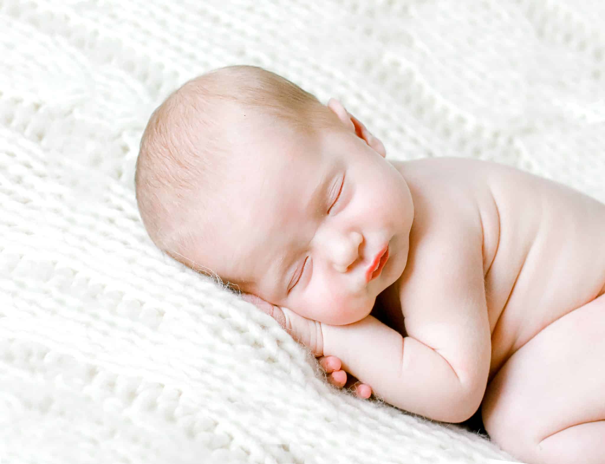 newborn baby laying on stomach faceing the camera sleeping on white blanket her hand under her head