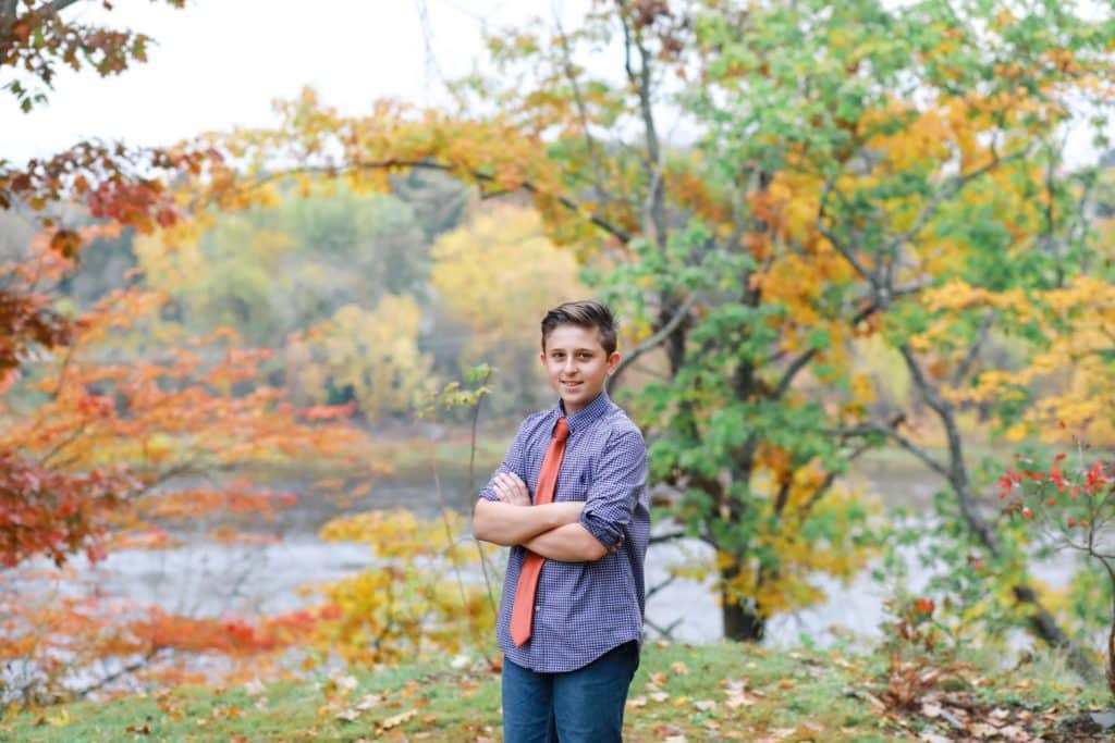 boy with a red tie adn plaid shirt stadning with arms crossed outside in the fall