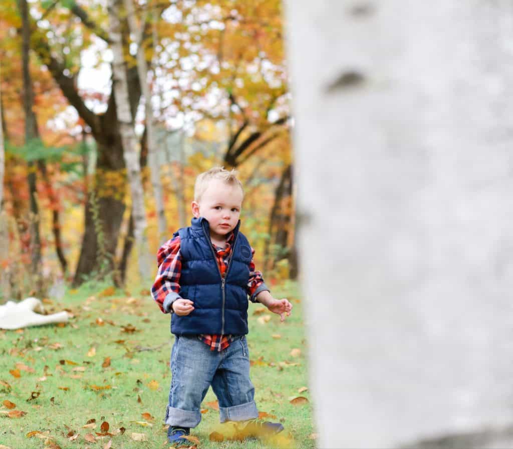 bangor maine toddler in plaid and a blue vest running around the yard in the fall behind a birch tree