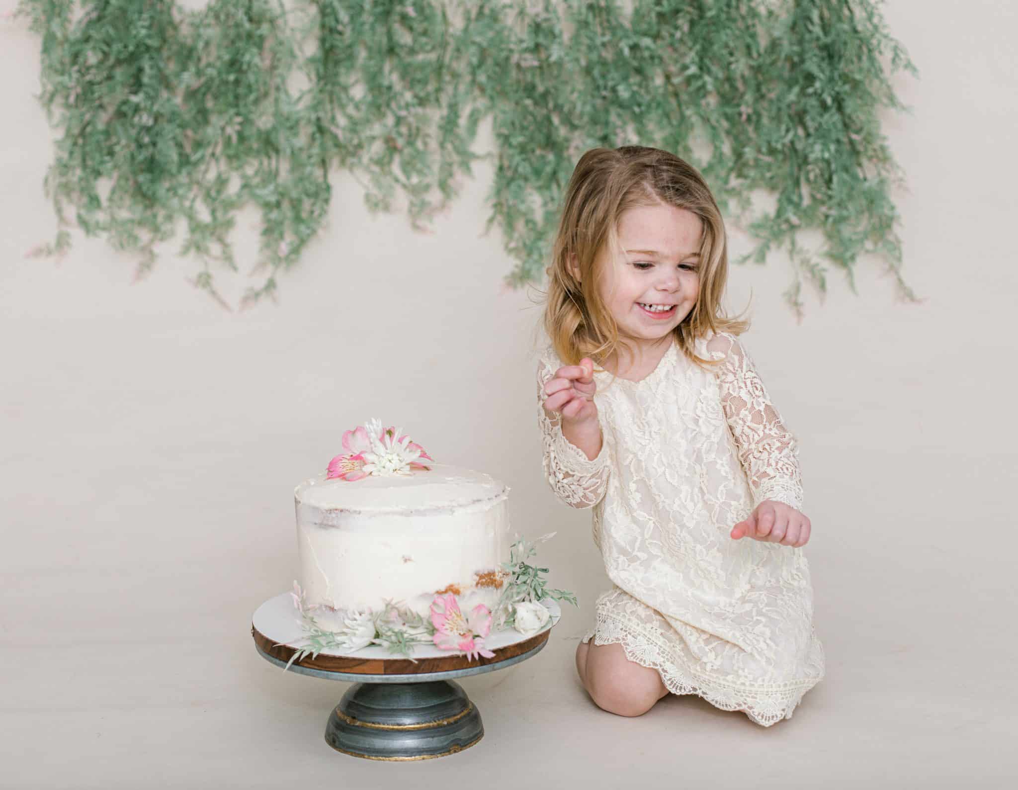 little girl laughing looking down, white cake, green garland, and white dress