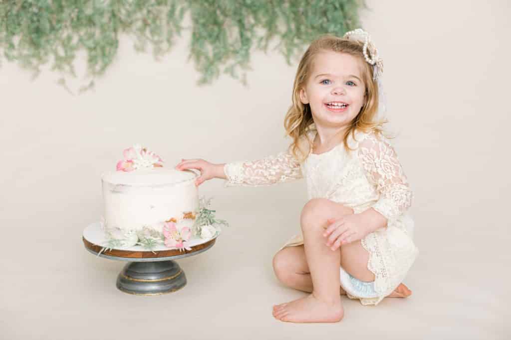 baby girl in a white dress with a botanical themed cake looking at the camera smiling 