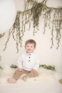 One year boy, white sweater, tan pants, white rug, green garland, smiling at the camera.