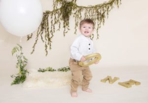 One year boy in a white sweater and tan pants smiling holding an o for one, green garland, large white balloon on a white fur rug, with a white background