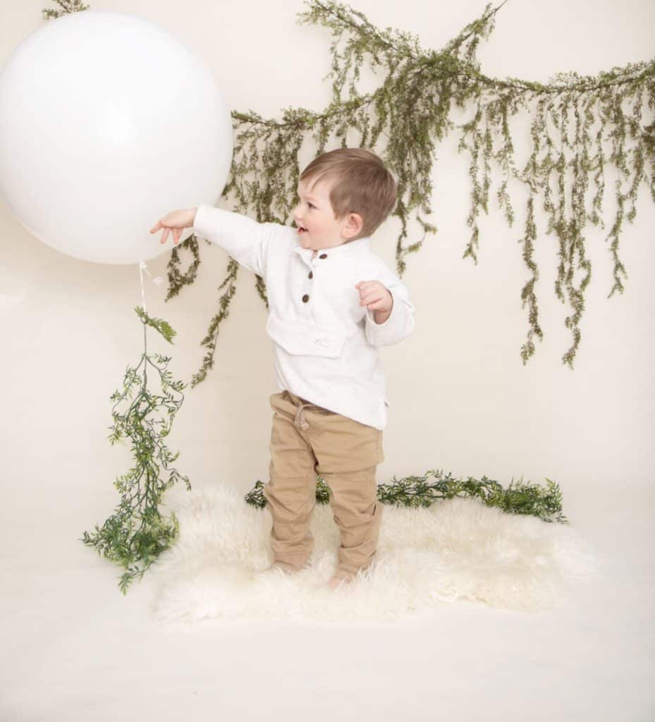 One year old boy in a white sweater and tan pants standing on a white rug, green garland, playing with a large white balloon 