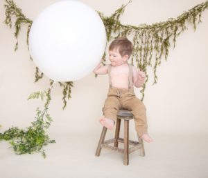 One year photos, large white balloon, baby boy in tan pants, suspenders sitting on a brown stool, with green garland.