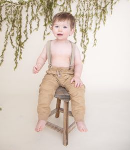 One Year boy sitting on a brown stool wearing suspenders and tan pants, green garland on an off white background.