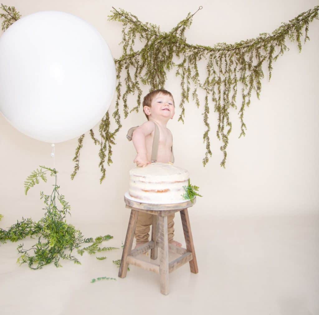 One year cake smash, large white balloon, green garland, off white background, one year old boy in suspenders, and tan pants