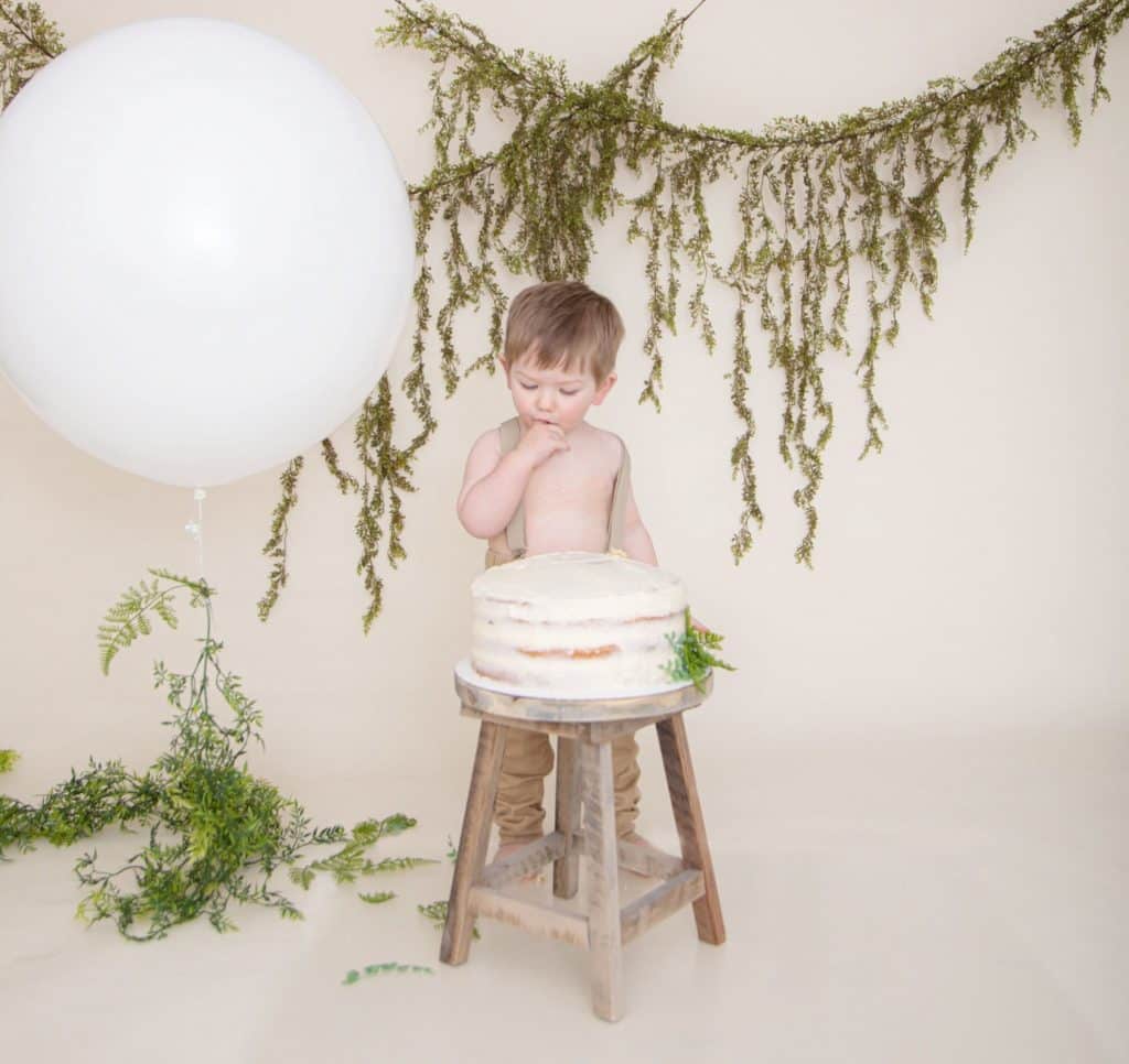 One year old boy in suspenders and tan pants eating a cake, white balloon, green garland, off white background