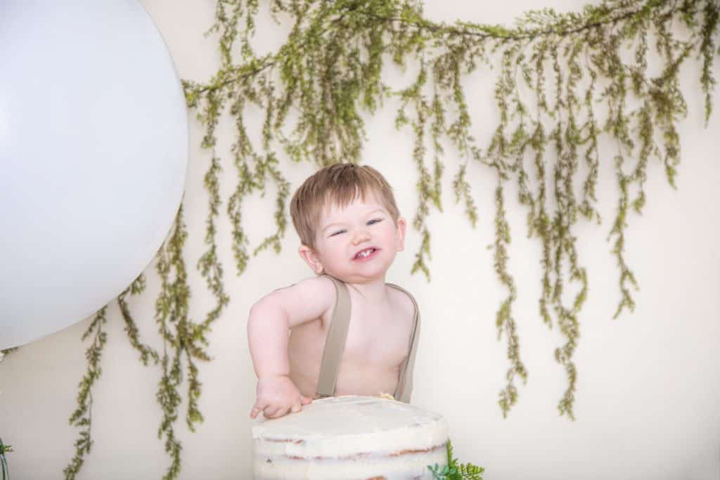 One year old boy, fingeer in the cake, green garland, silly smiling at the camera