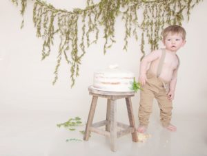 One year old boy in suspenders and tan pants, cake smash, green garland, large white balloon, fur