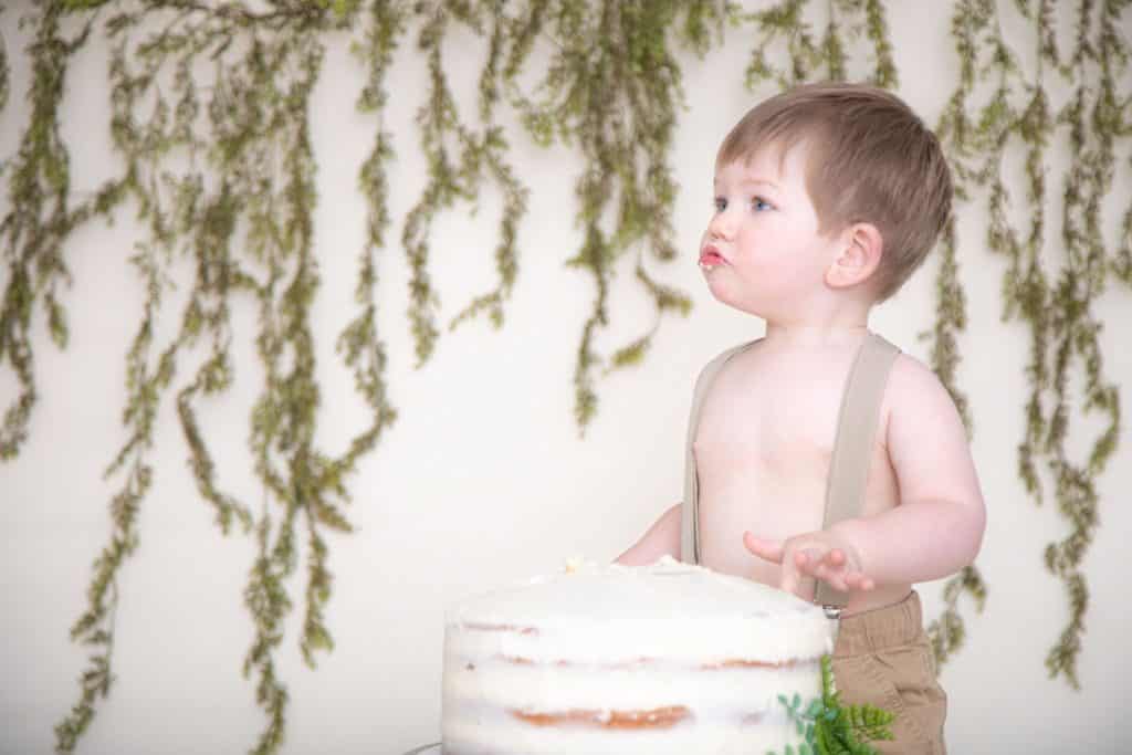 One Year Old Boy in suspenders and tan pants, green garland, no shirt, silly face