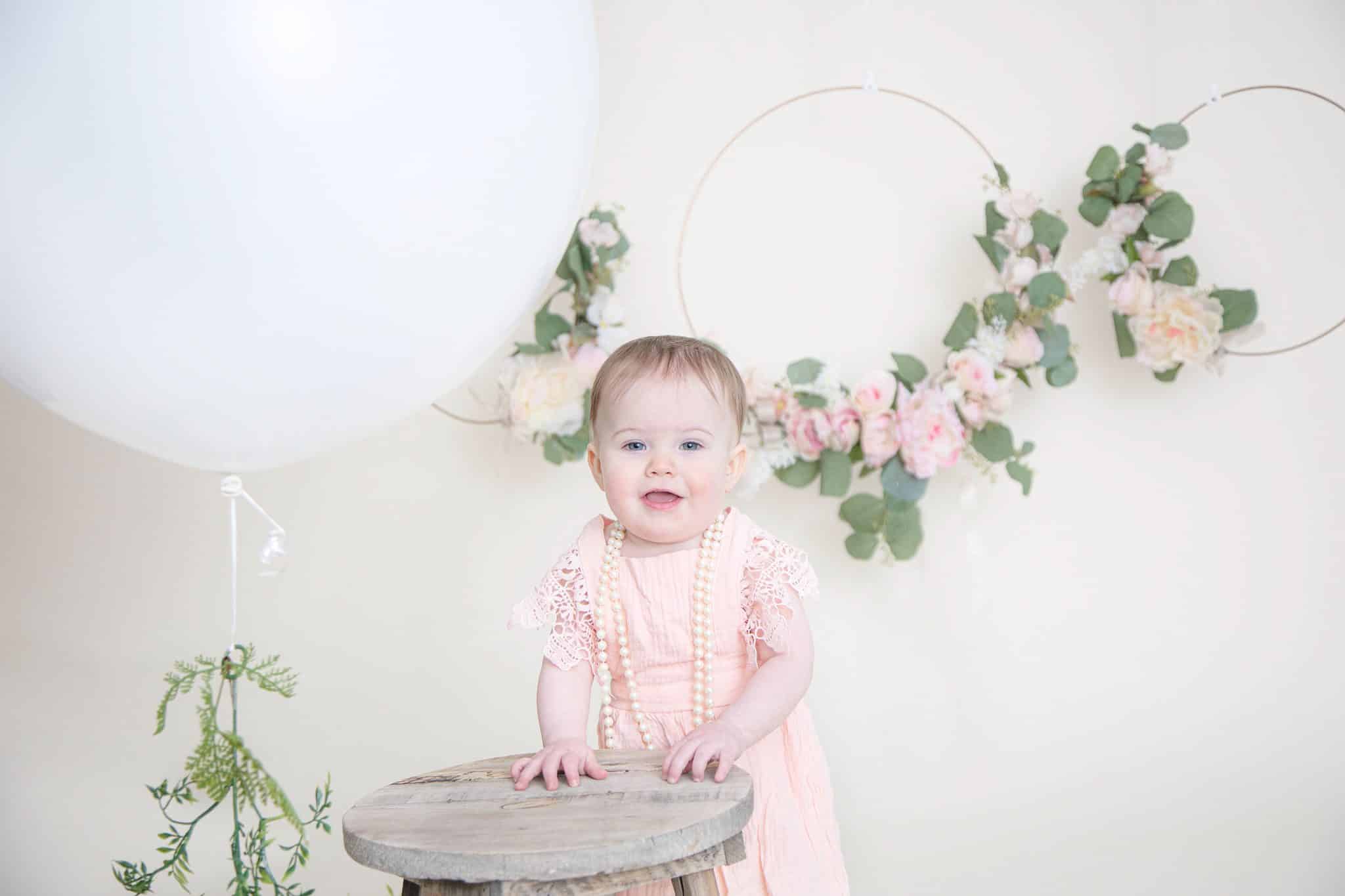 one year old in pink dress hands on a wooden stool smiling at the camera