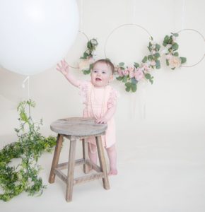 One year girl looking at the camera smiling reaching to touch a large white balloon holding onto the brown stool