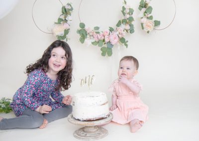 One year old and sister looking at the camera and eating cake