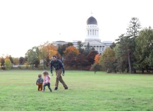 dad chasing his two little kids in the grass near the augusta, MAine captiol