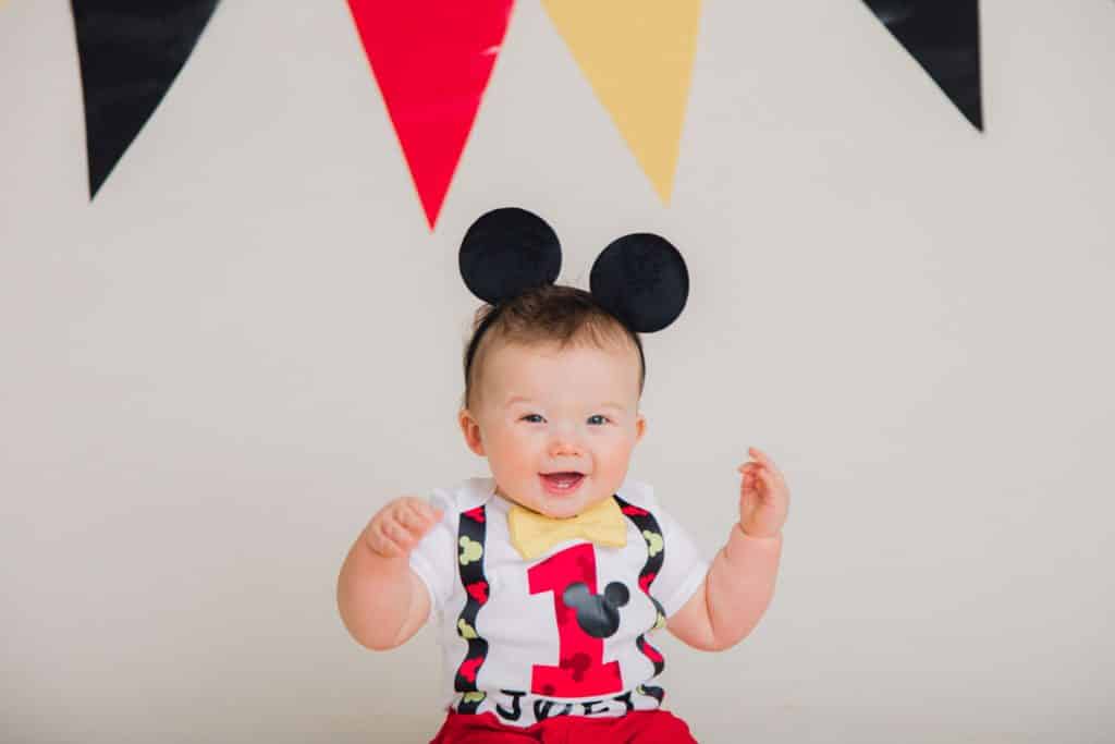 red, yellow, black banner, one year old with mickey mouse ears smiling and looking at the camera with a red one on his shrt