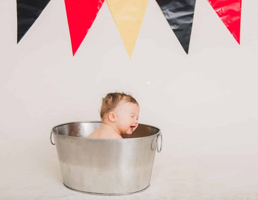 red, yellow, black banner in the background. One year boy in a silver bucket with bubbles smiling