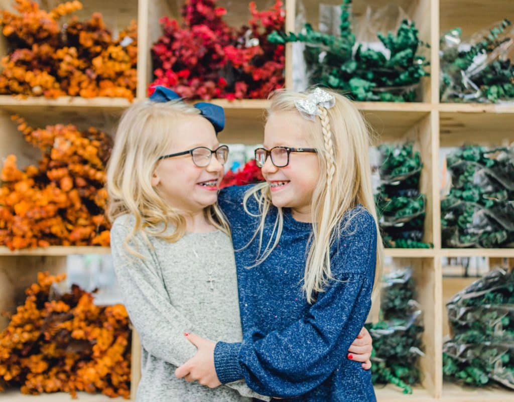 twin blonde girls with glasses holding each other and looking at each other with colors behind them