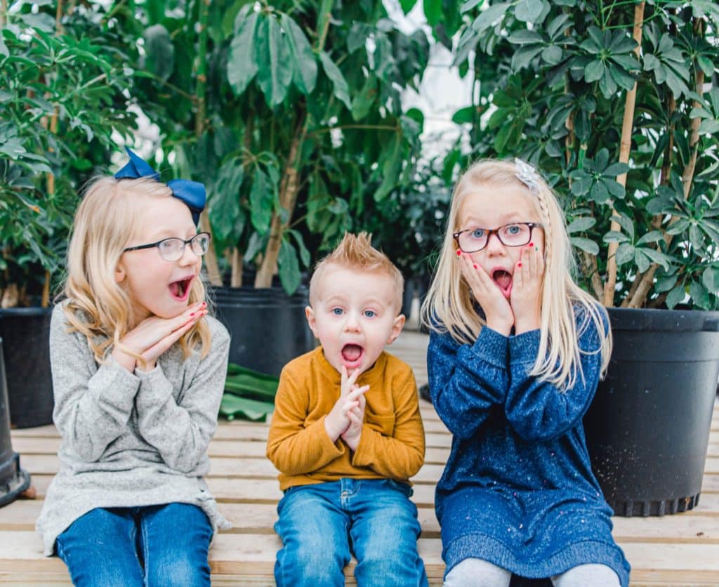 three kids. Two girls and one boy making a silly face in a greenhouse 